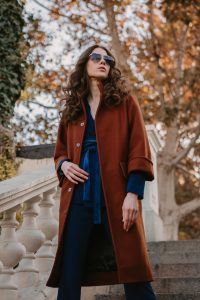 beautiful stylish smiling skinny woman with curly hair walking in street stairs dressed in warm brown coat and blue suit, autumn trendy fashion street style, accessories sunglasses